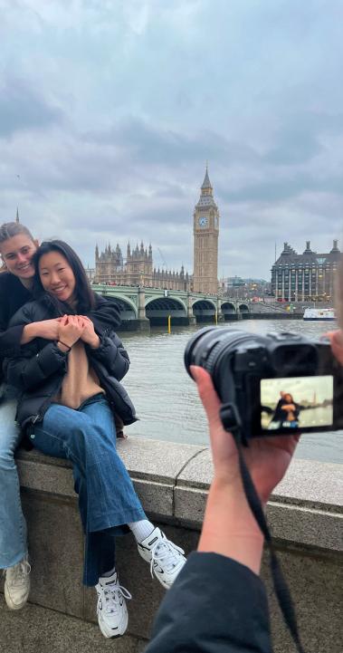 two students sitting on ledge hugging while another student holds up a camera and takes a picture