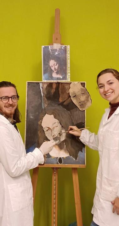 two students in white coats holding paint brushes in front of a painting on an aisle